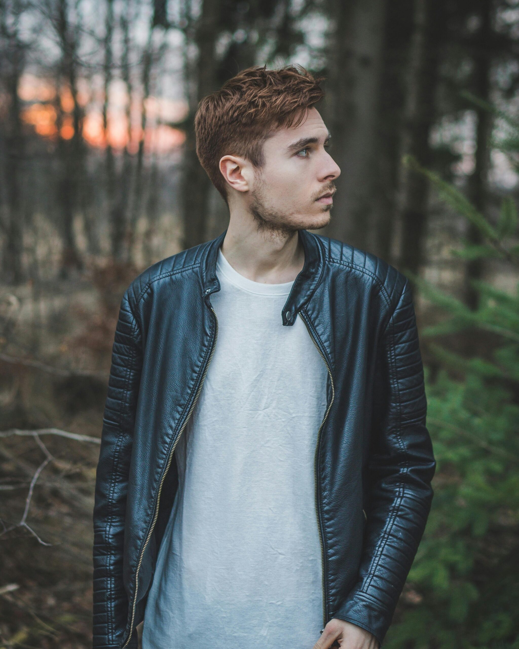 Young man in a black leather jacket standing in a forest during twilight, styled fashion portrait.